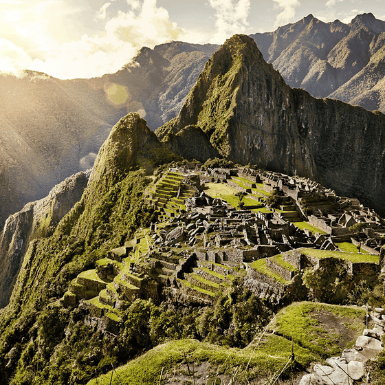 Sunlit Machu Picchu with stone ruins set against lush green mountains under a partly cloudy sky.