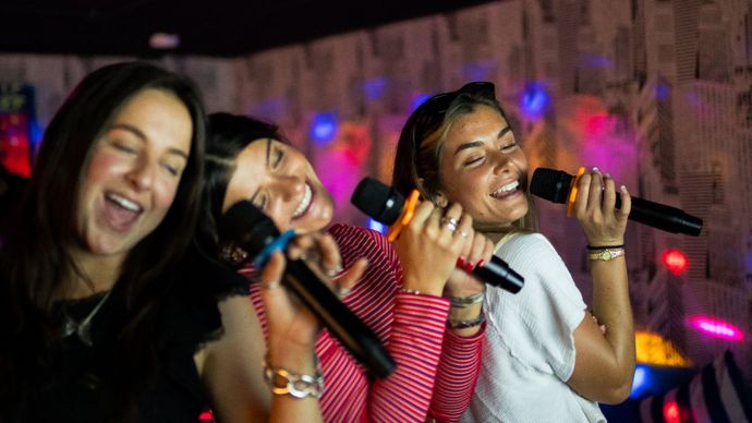 Three women joyfully singing into microphones at a karaoke session, with colorful lights in the background.
