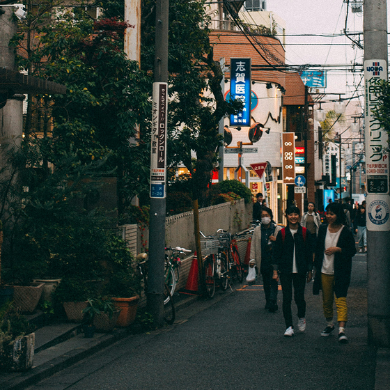A narrow urban street with people walking, bicycles parked, greenery, and colorful signs in an atmospheric evening light.
