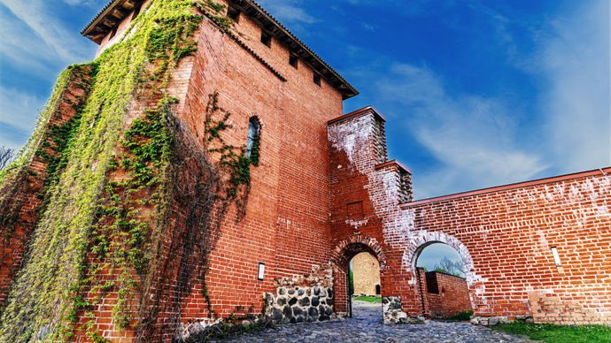 A historic red brick tower with ivy climbing the facade, connected to an arched entrance, under a blue sky with scattered clouds.