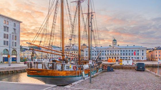 A vintage sailing ship docked at a harbor with a historic building and a cloudy sunset sky in the background.