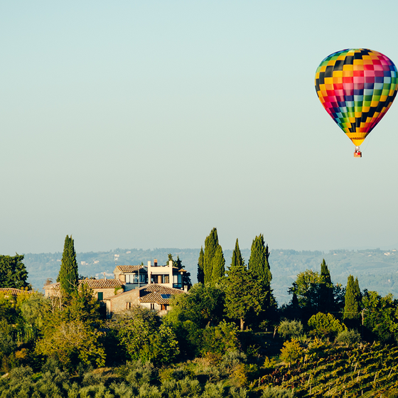 A colorful hot air balloon floats over a scenic landscape with a hillside villa surrounded by trees and vineyards under a clear blue sky.