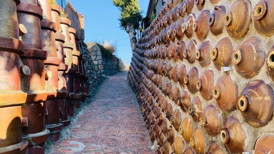 Narrow path between walls made of stacked clay pots, with patterned pavement and a clear blue sky above.