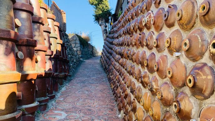 Narrow path between walls made of stacked clay pots, with patterned pavement and a clear blue sky above.