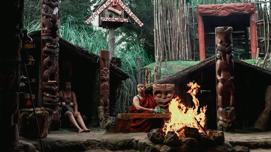 Two people in traditional attire sit near a fire, surrounded by carved wooden structures in a lush, forested setting.