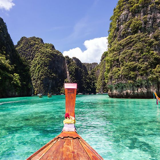 A traditional wooden boat floats on clear turquoise water surrounded by tall, lush limestone cliffs under a bright blue sky.