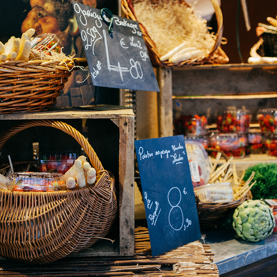 Rustic market display with wicker baskets of white asparagus, jars of preserves, artichokes, and chalkboard price signs.