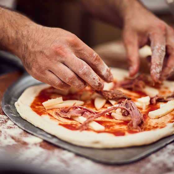 Hands preparing a pizza with tomato sauce, cheese, and meat on a floured countertop, ready for baking.