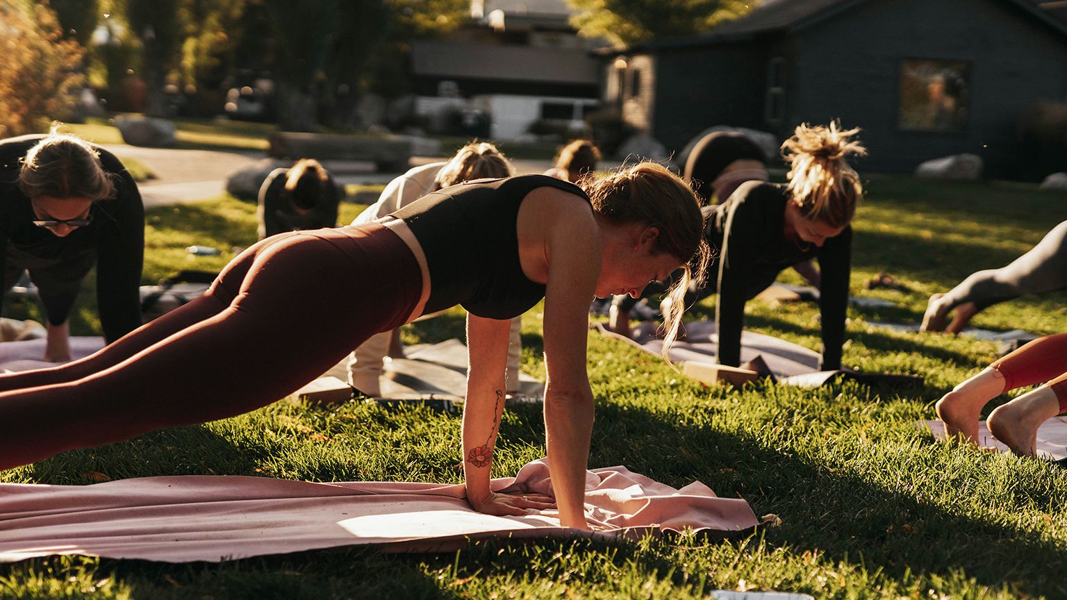 People practicing yoga outdoors on a sunny day, performing a plank pose on yoga mats spread across the grass.