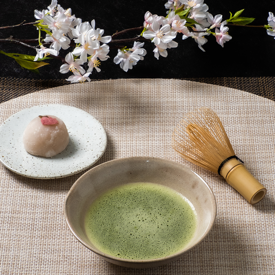 A bowl of matcha tea with a bamboo whisk, a sweet pastry on a plate, and cherry blossoms on a woven mat.