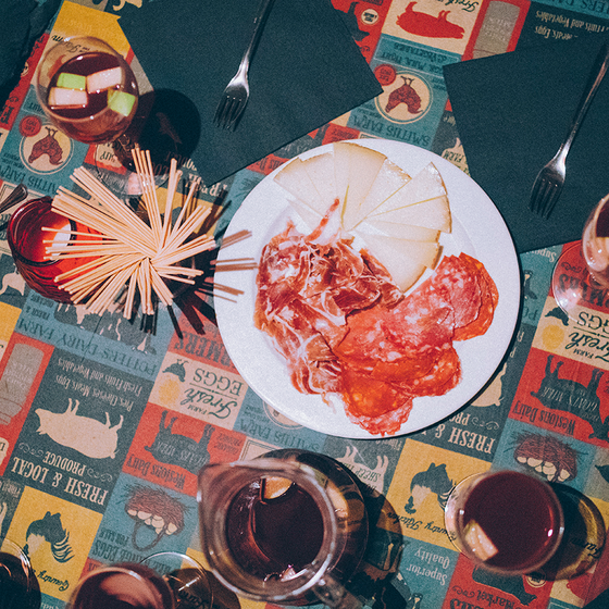 A vibrant table setting with a platter of cured meats and cheese, surrounded by drinks, bread, and forks on a colorful, patterned tablecloth.
