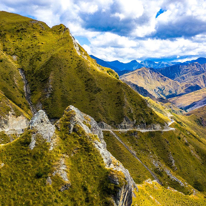 A winding road cuts through rugged, green hills under a blue sky with scattered clouds, with distant mountains in the background.