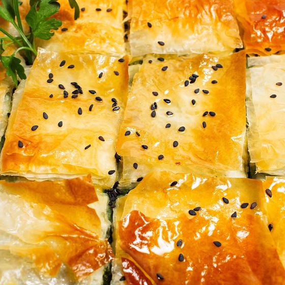 Close-up of golden, flaky pastry squares topped with black sesame seeds, garnished with a sprig of fresh parsley.