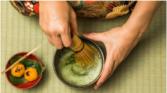Hands preparing matcha tea with a bamboo whisk on a tatami mat, next to a small plate with two yellow sweets on a leaf.