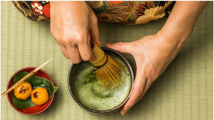Hands preparing matcha tea with a bamboo whisk on a tatami mat, next to a small plate with two yellow sweets on a leaf.