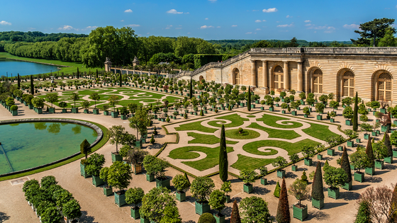An ornate garden with manicured lawns, geometric patterns, and potted trees, adjacent to a grand building with arched windows and columns.