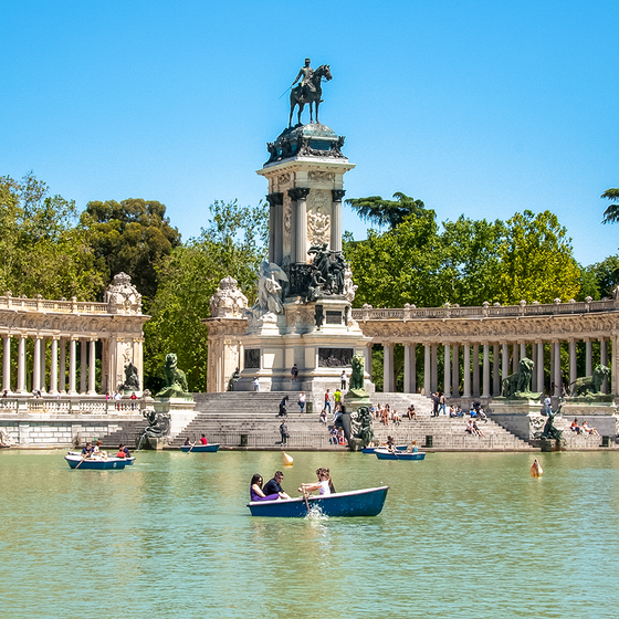 People rowing boats on a lake in front of the Alfonso XII monument in Madrid's Retiro Park, with clear blue skies and lush trees in the background.