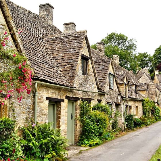 Quaint stone cottages with steep roofs and flowering vines line a narrow, lush green lane in a picturesque village setting.