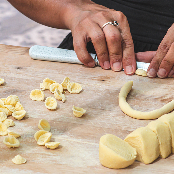 Hands shaping pasta dough into orecchiette on a wooden surface, with dough pieces and a rolling pin visible.