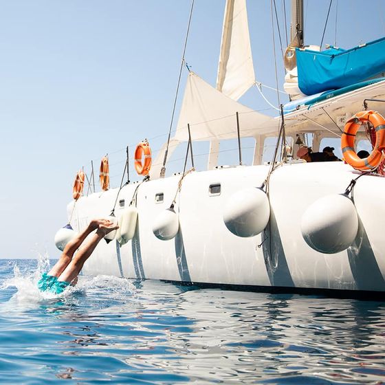 Person diving into the sea from a yacht, with a rocky coastline in the background under a clear blue sky.