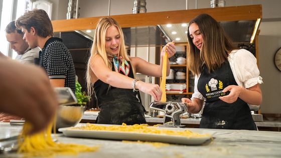 Two women making fresh pasta using a pasta machine in a bright kitchen, with others engaged in cooking activities in the background.