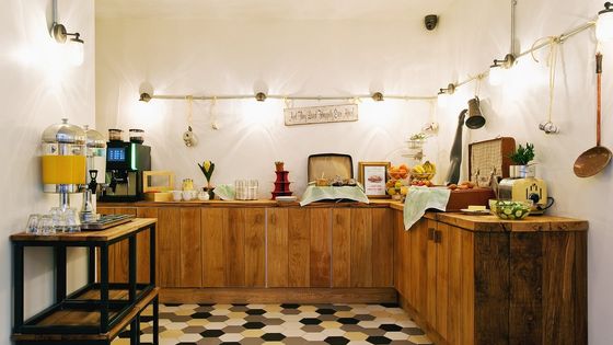 Rustic breakfast buffet with wooden cabinets, tea and coffee dispensers, bread, fruits, and decorative lights on a tiled floor.