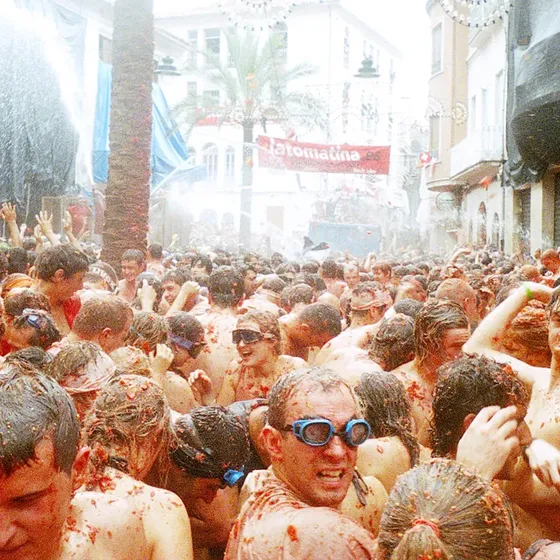A large crowd of people covered in tomatoes participates in the Tomatina festival, with some wearing goggles, amidst a street filled with smashed tomatoes.