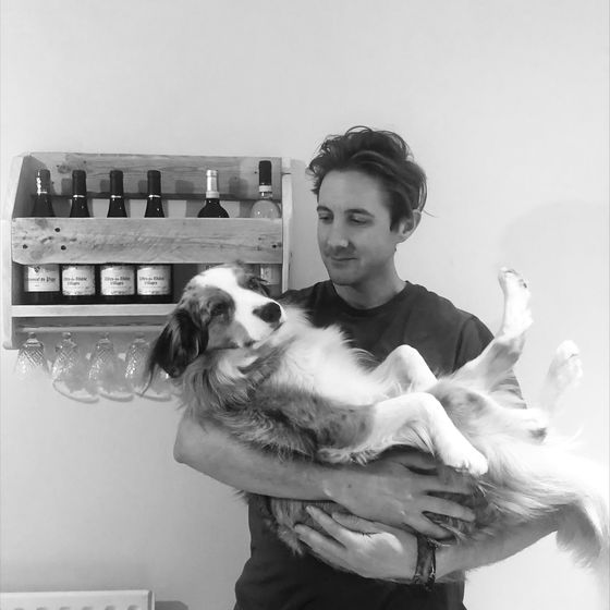Man holding a relaxed dog indoors, with a wine rack and glasses on the wall in the background. Black and white image.