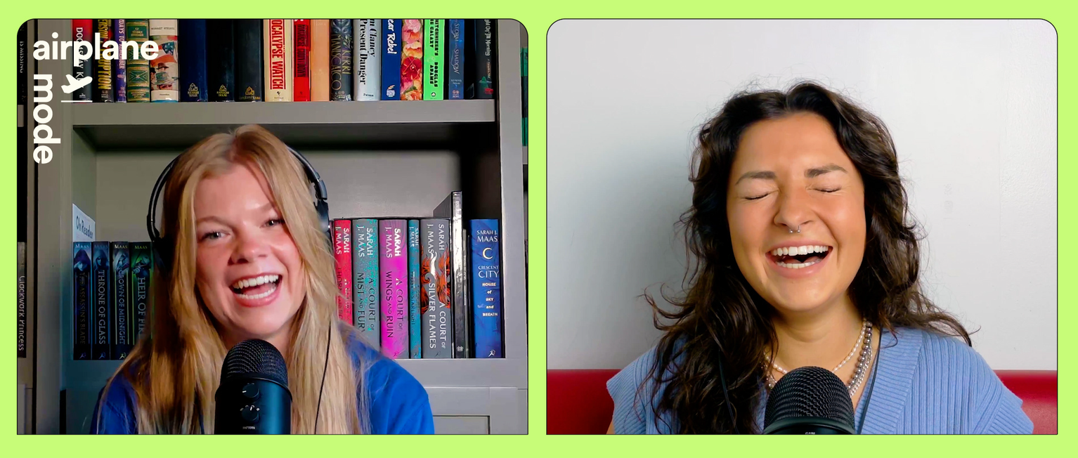 Two women with microphones laugh during a recording session. One sits in front of bookshelves, the other against a white wall.