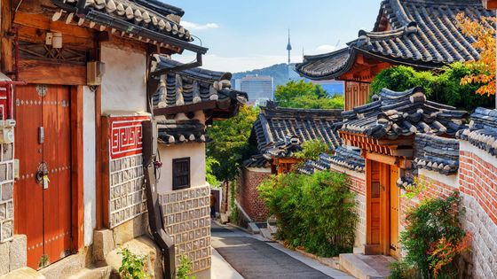 Traditional Korean hanok village with narrow street, tiled roofs, brick walls, and a tower in the distance under a clear blue sky.