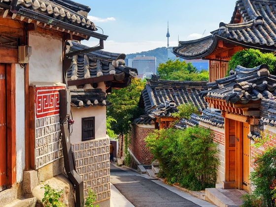 Traditional Korean hanok village with narrow street, tiled roofs, brick walls, and a tower in the distance under a clear blue sky.