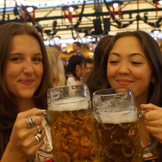 Two people at a festive event smiling and toasting with large mugs of beer, surrounded by a lively crowd and festive decorations.