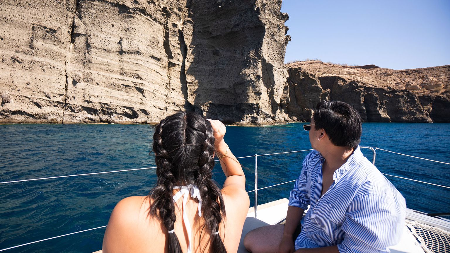Two people on a sailboat, one with braided hair and one in sunglasses, looking at tall eroded cliffs above deep blue water.