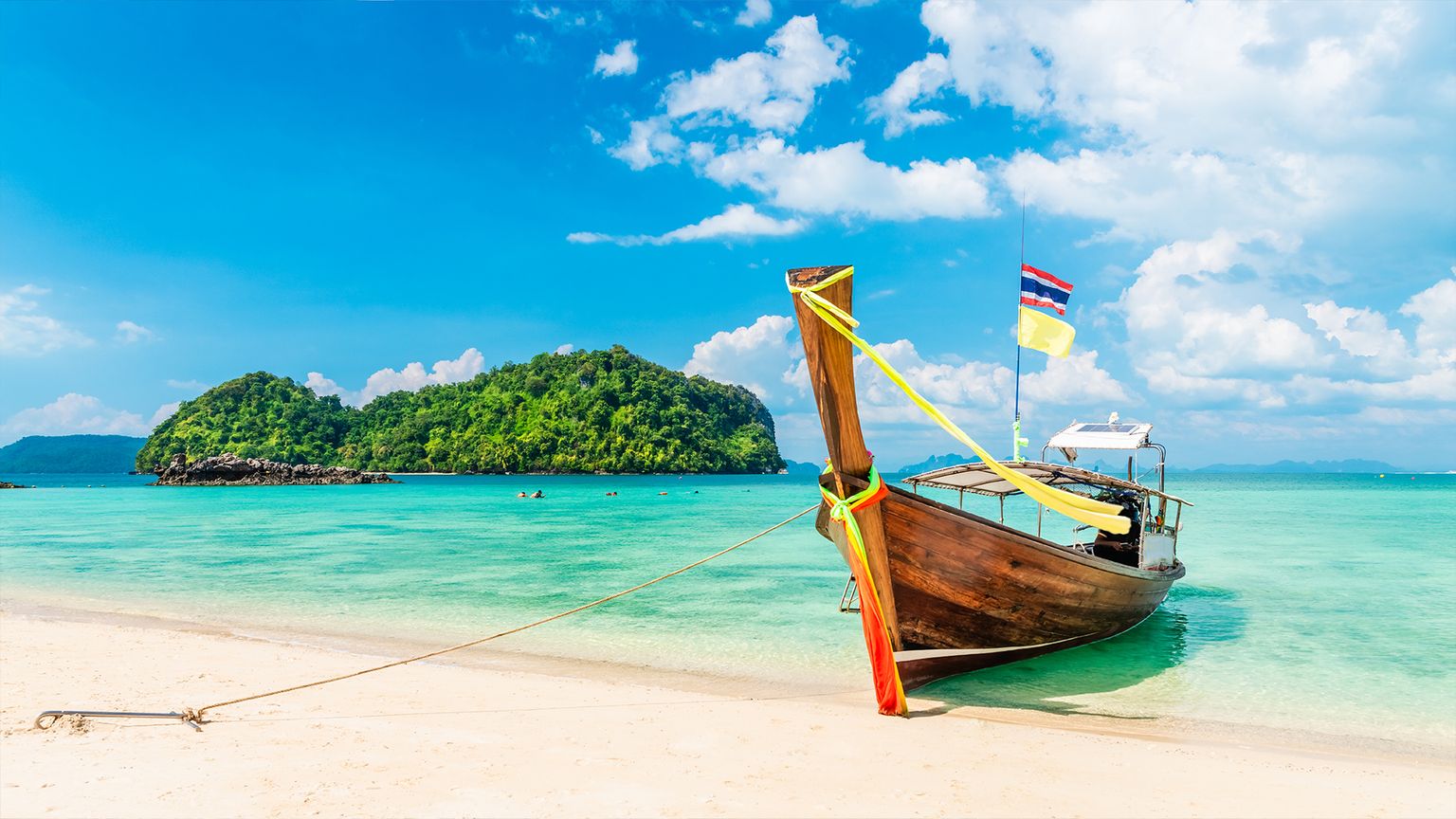 A wooden boat with flags is moored on a sandy beach with turquoise water and lush green islands under a blue sky with clouds.