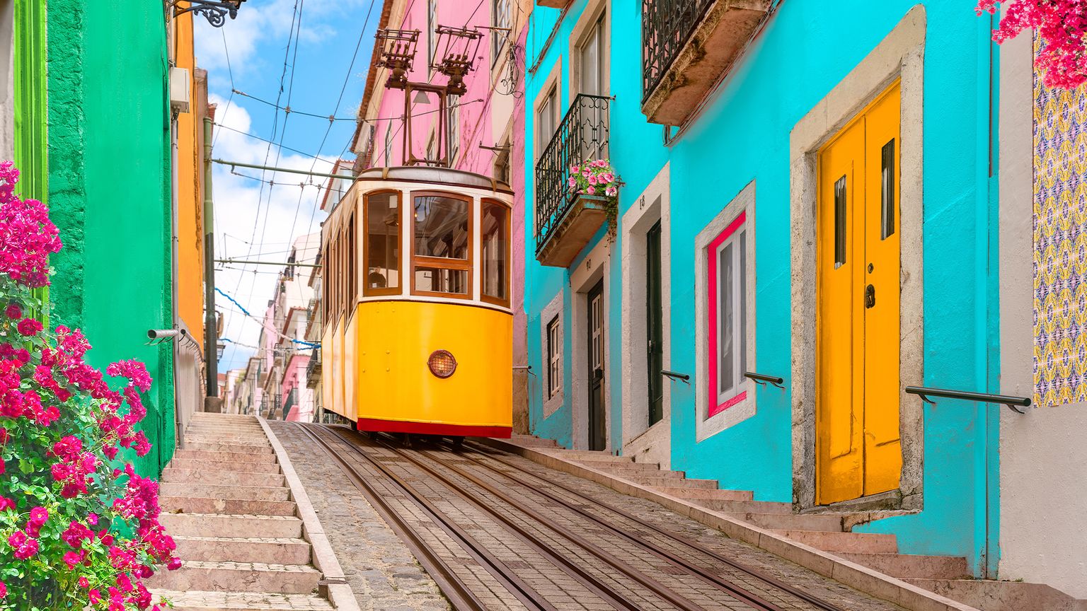 A vibrant yellow tram ascends a colorful street with pink, blue, and green buildings, adorned with pink flowers on a sunny day.