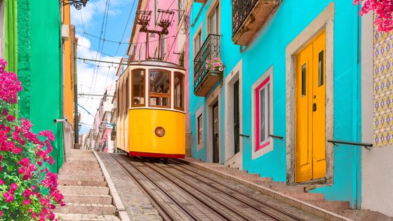 A vibrant yellow tram ascends a colorful street with pink, blue, and green buildings, adorned with pink flowers on a sunny day.