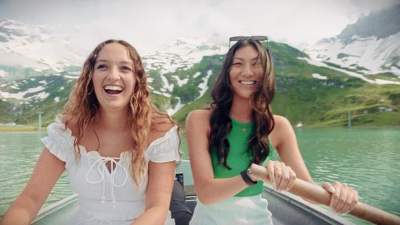Two women smiling in a rowboat on a lake, surrounded by snow-capped mountains and green hills under a cloudy sky.