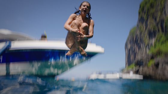 Person mid-air doing a cannonball into the ocean, with a blurred boat and rocky cliff in the background, under a clear blue sky.