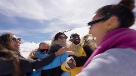 A group of people in winter clothing laughing and playing in the snow on a sunny day, with mountains in the background.