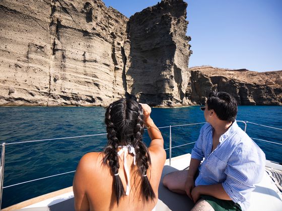 A man and woman sit on a boat, gazing at towering rocky cliffs under a clear blue sky.