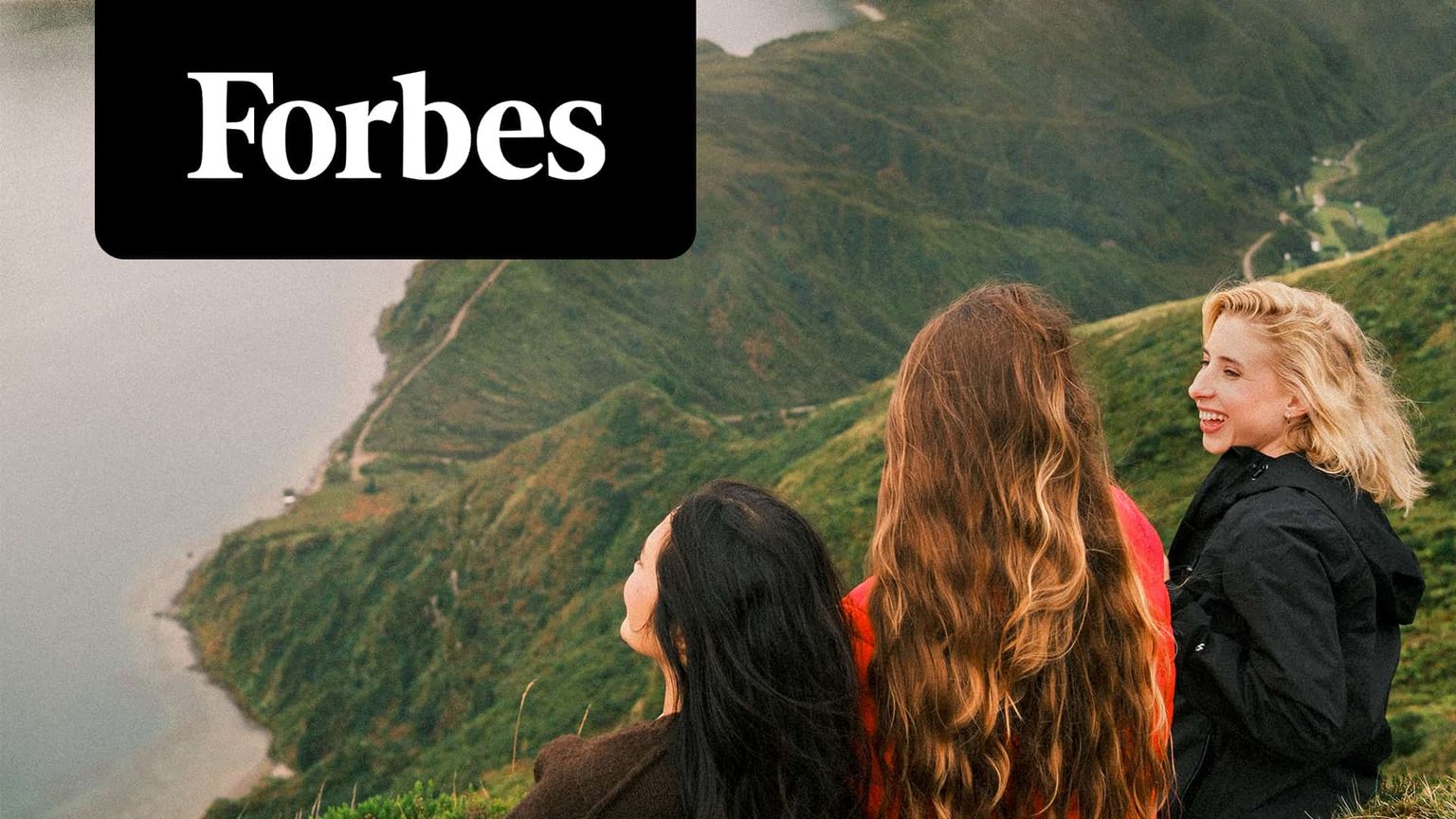 Three women overlook a scenic landscape from a hilltop, with lush greenery and a winding coastline. The Forbes logo is in the top left corner.