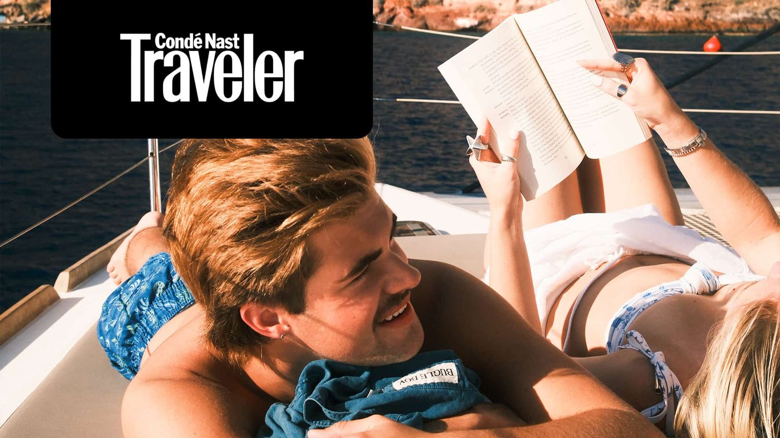 Two people relaxing on a boat under the sun. One reads a book while the other smiles, with water and rocky coastline in the background.