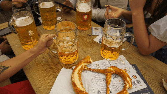 People at a wooden table with large beer mugs and pretzels, one person holding a mug. Various cards and papers are scattered on the table.