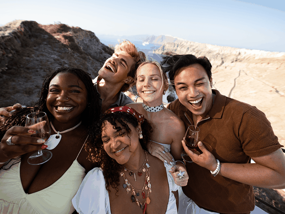 Five friends joyfully smile and pose with drinks on a sunny, rocky coastal overlook, capturing a moment of celebration and happiness.
