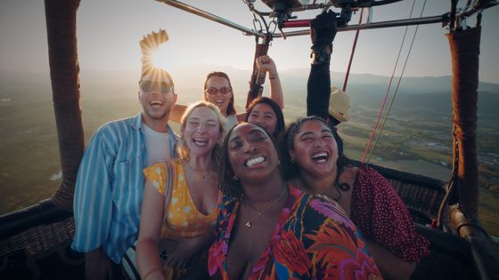 A joyful group of six people smiles and poses for a selfie in a hot air balloon during sunset, with scenic mountains in the background.