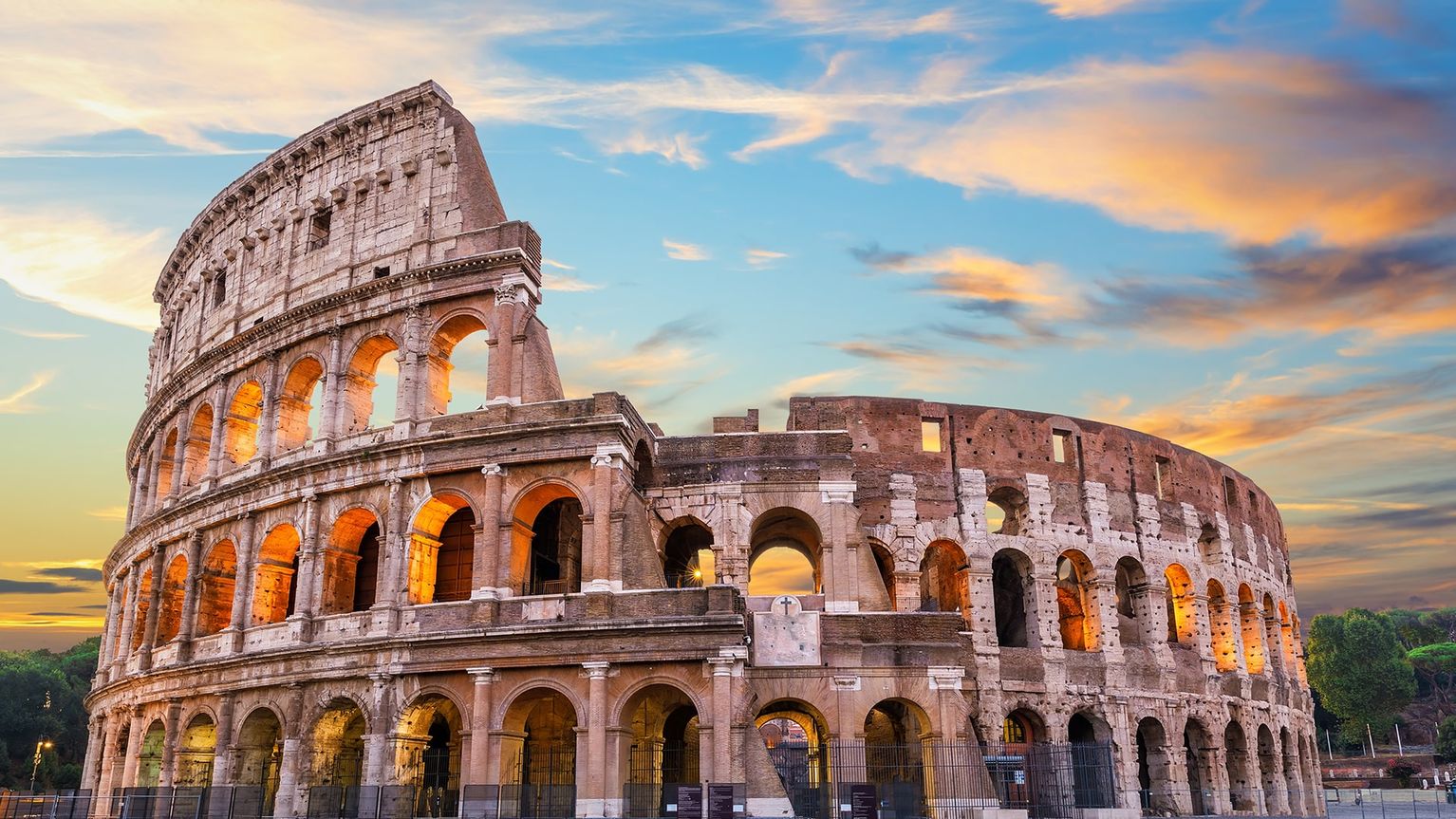 The Colosseum bathed in golden light at sunset, with a vibrant sky of orange, pink, and blue hues in the background.