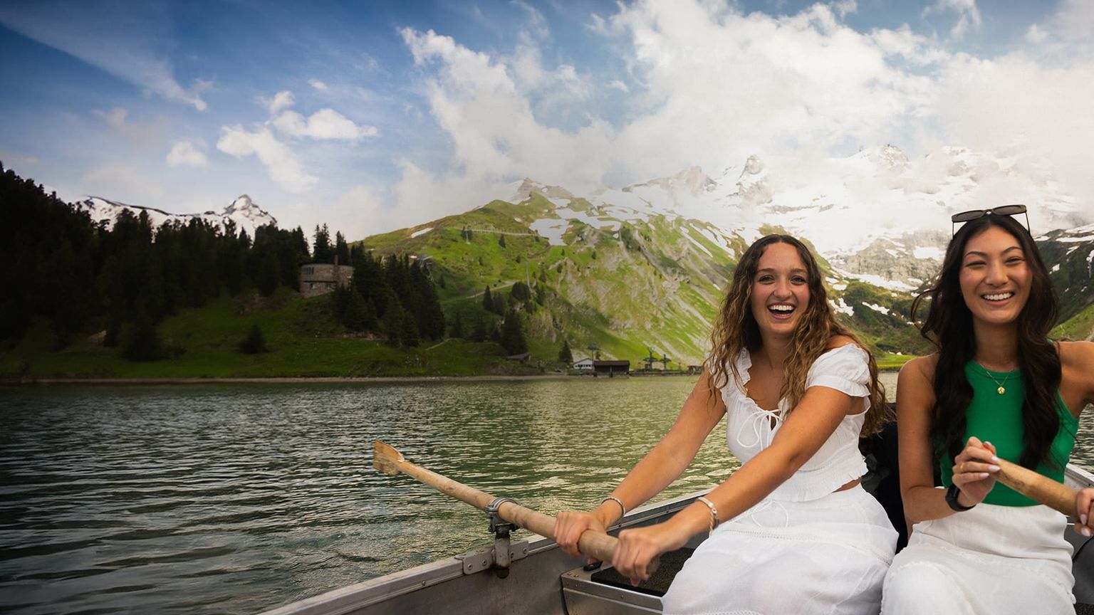 Two women smiling and rowing a boat on a serene lake, with lush green hills and snow-capped mountains in the background.