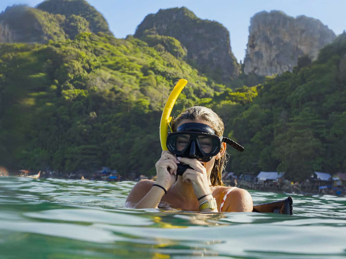 A person wearing a snorkel mask is in the clear water, with lush green hills and rocky cliffs in the background.