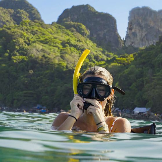 A person wearing a snorkel mask is in the clear water, with lush green hills and rocky cliffs in the background.