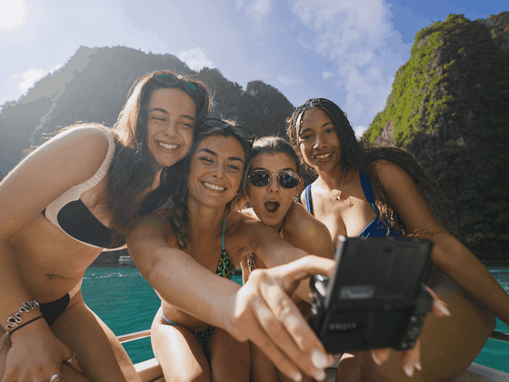 Four women in swimsuits pose for a selfie on a boat, surrounded by clear blue water and lush green cliffs.
